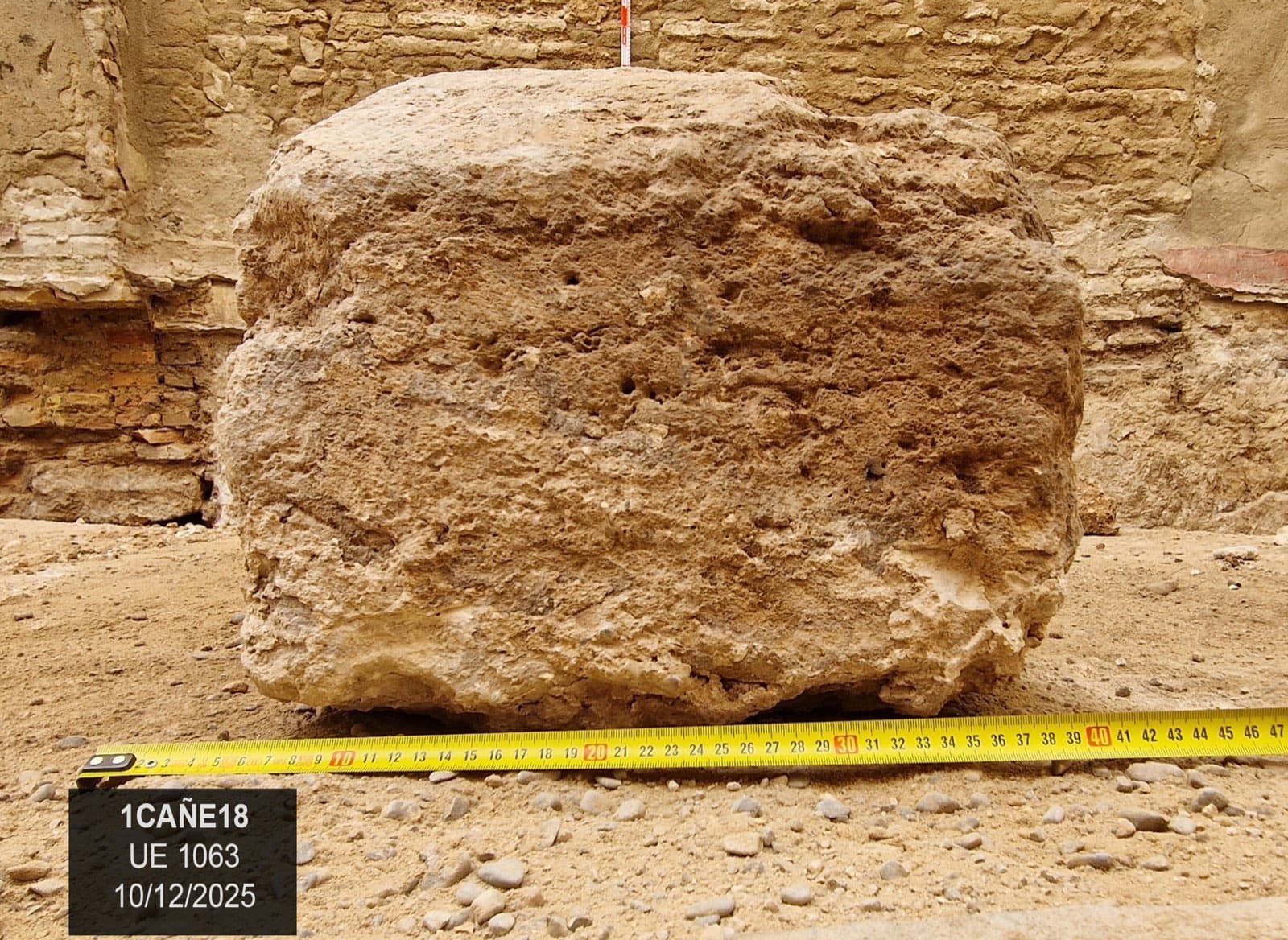 Een bewerkt stenen blok, van boven afgebroken, tegen een muur, met een gele rolmaat bij de voet en het bordje van de opgraving (“1CAÑE18, UE 1063, 10/12/2025”).