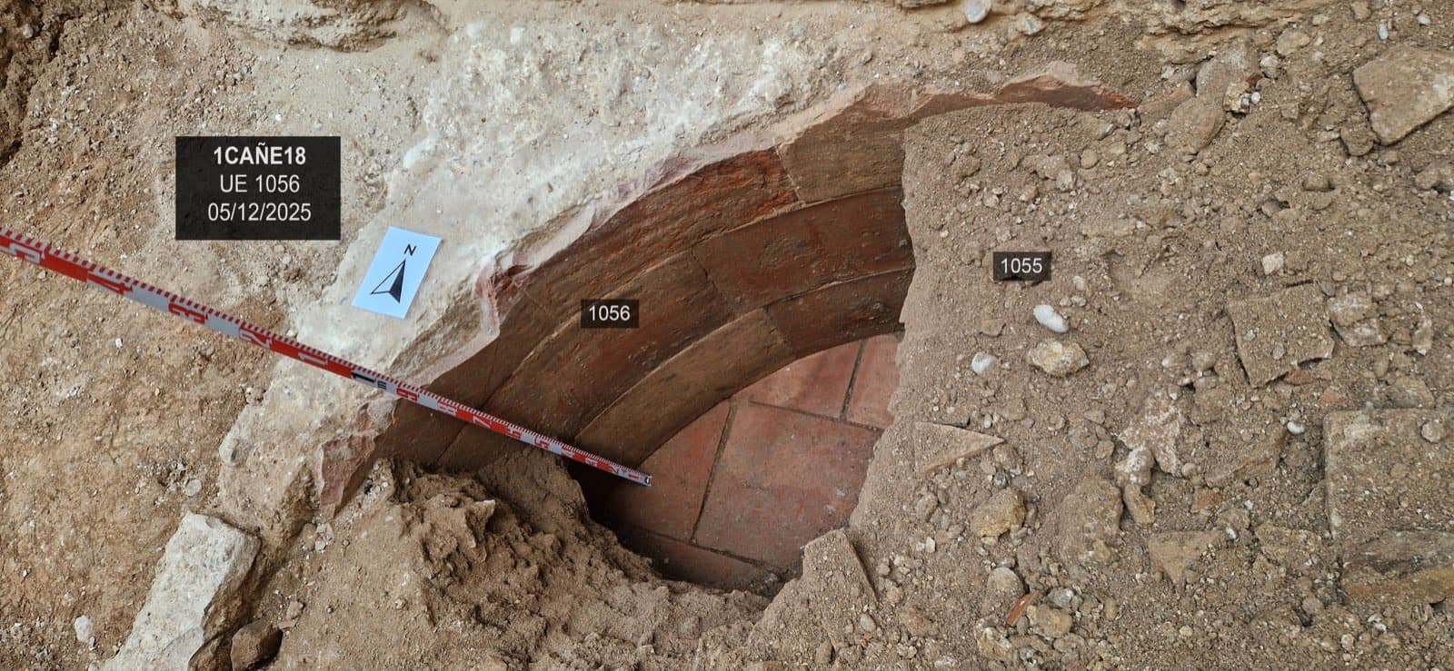 De gebogen rand van een structuur bekleed met rode baksteen, van bovenaf gezien, met een schaallat diagonaal erover, een noordpijl en het bordje van de opgraving (“1CAÑE18, UE 1056, 05/12/2025”).