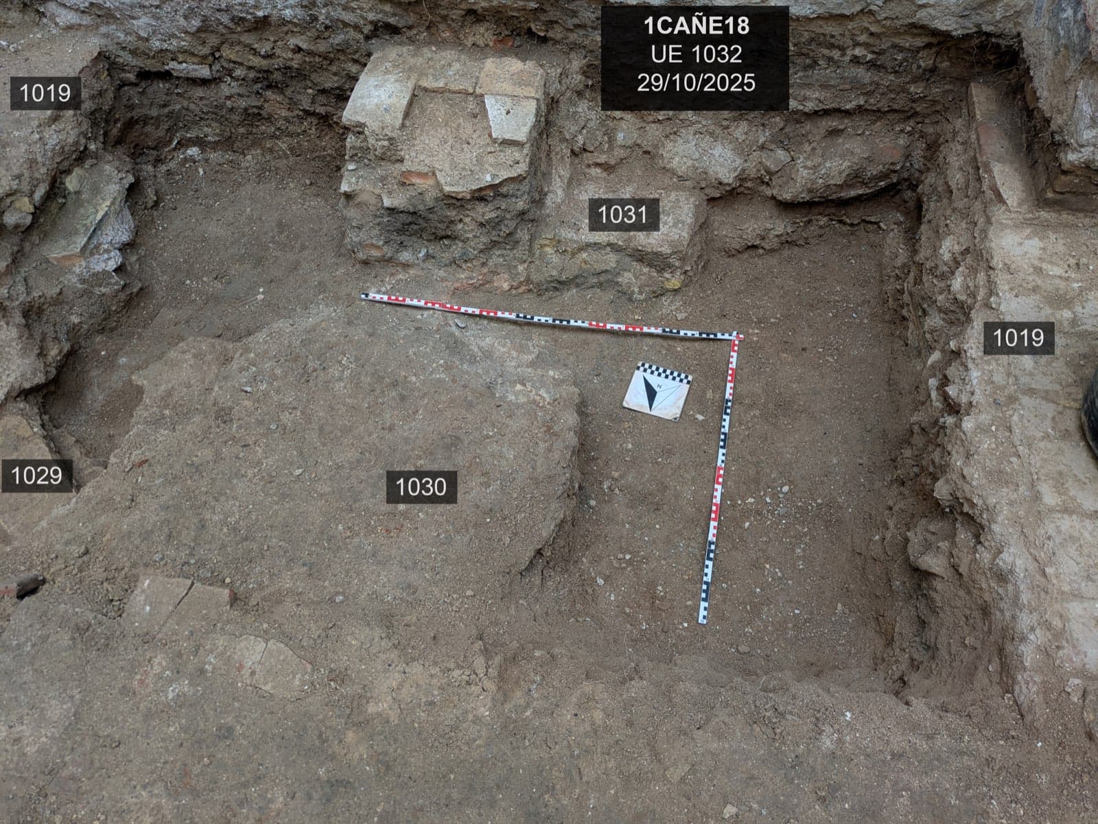 Een vierkante opgegraven kuil in de grond, met het bordje van de opgraving (“1CAÑE18, UE 1032, 29/10/2025”), een noordpijl en een schaallat, naast de nummers van verschillende stratigrafische eenheden op de grond.