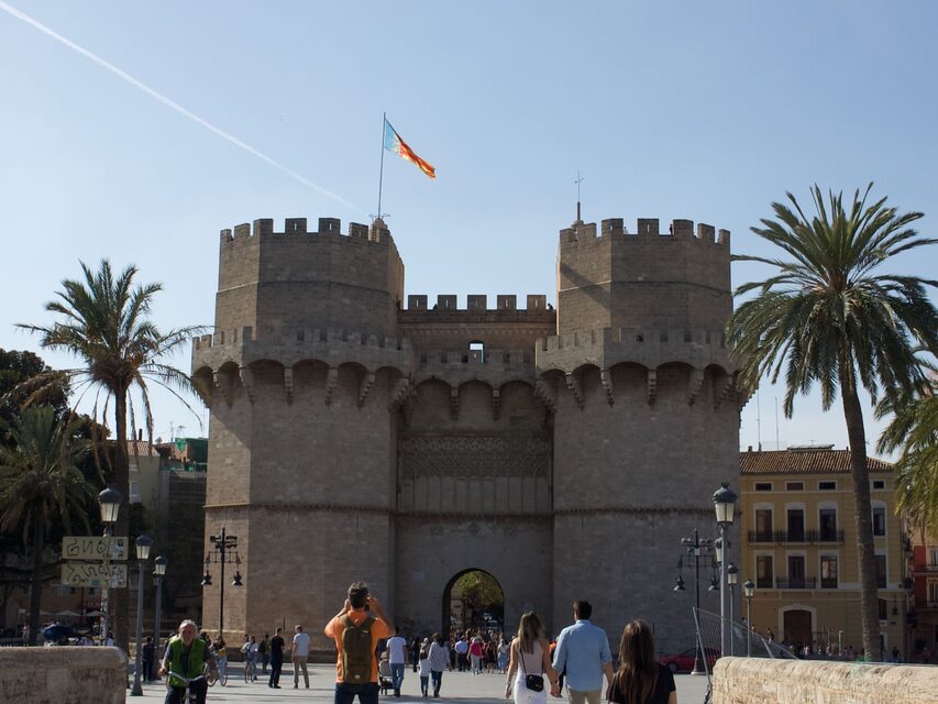 De Torres de Serranos gezien vanaf de brug, met de hele poort, de vlag van de Valenciaanse Gemeenschap en palmbomen aan weerszijden.