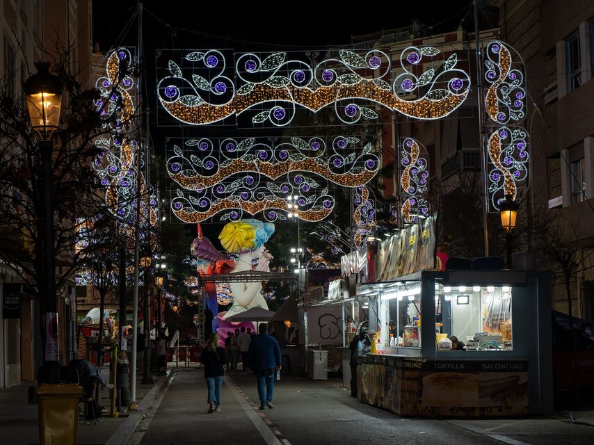 Een straat verlicht met lichtbogen tijdens de Fallas, met een falla op de achtergrond en eetkraampjes rechts.