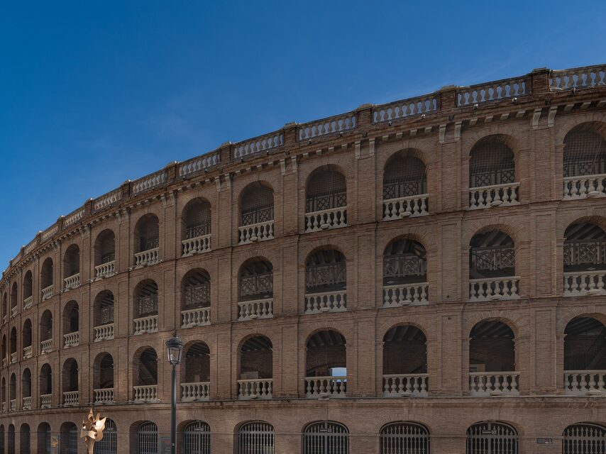De gebogen bakstenen gevel van de Plaza de Toros, met rijen bogen en witte balustrades onder een blauwe lucht.